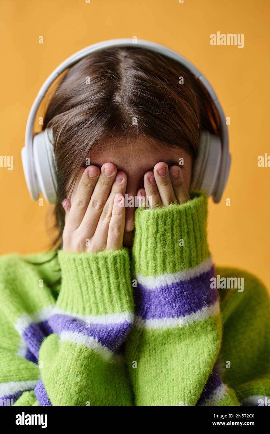 Porträt eines Mädchens im Teenageralter, das ihr Gesicht mit den Händen bedeckt, während es Musik über Kopfhörer hört, isoliert auf orangefarbenem Hintergrund Stockfoto
