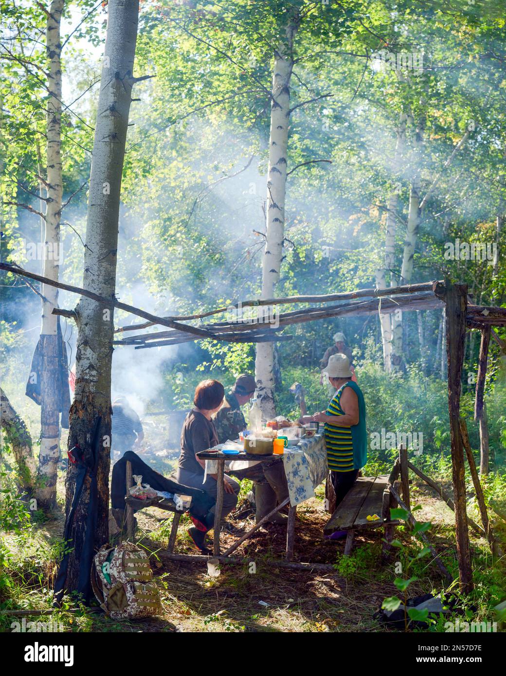 Ältere Yakuts decken den Tisch an einem Ort, um sich im wilden nördlichen Wald im Rauch des Feuers in den Sonnenstrahlen, die durch den Baum fallen, auszuruhen Stockfoto