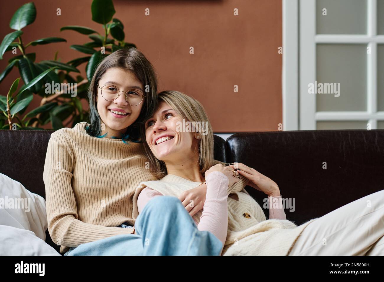 Glückliches Teenager Mädchen umarmte ihre Mutter und lächelte, während sie sich auf dem Sofa im Zimmer ausruhten Stockfoto