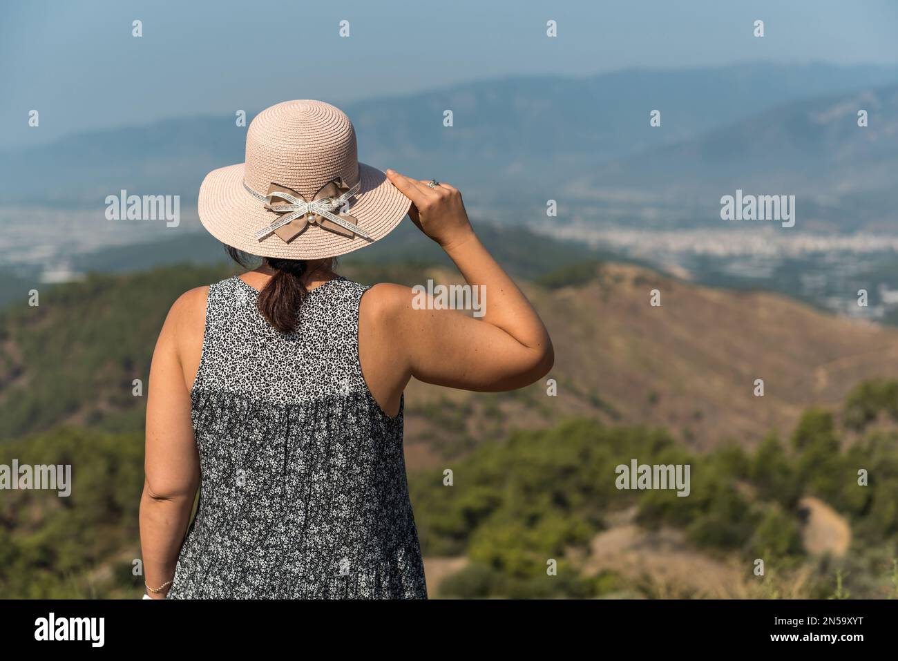 Junge Frau blickt in die Ferne, wunderschöne Berge im Hintergrund, denkt tief nach, trägt ein Kleid und einen Hut, Natur, langsames Leben, Reisen Stockfoto