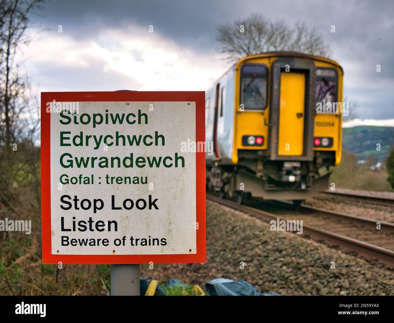 Ein Zug passiert ein Warnschild mit der Aufschrift „Stop Look Listen“ auf Walisisch und Englisch an einem Fußweg, der eine Bahnlinie kreuzt. An einem bedeckten Tag im Winter Stockfoto