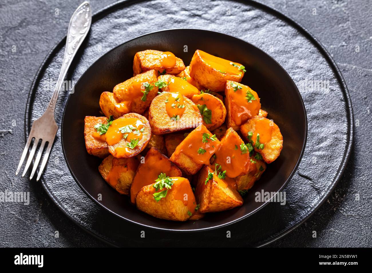 Patatas bravas, frittierte Kartoffeln mit scharfer Sauce in schwarzer Schüssel auf Betontisch, spanische Küche Stockfoto