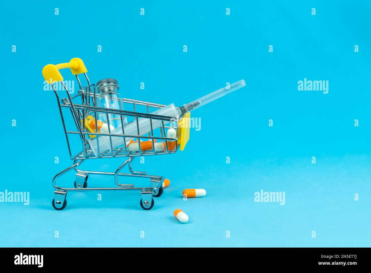Einkaufswagen mit medizinischer Ampulle, Spritze und Kapseln auf blauem Hintergrund, Kopierraum. Konzept des Arzneimittelkaufs, Erhöhung der Arzneimittelpreise Stockfoto