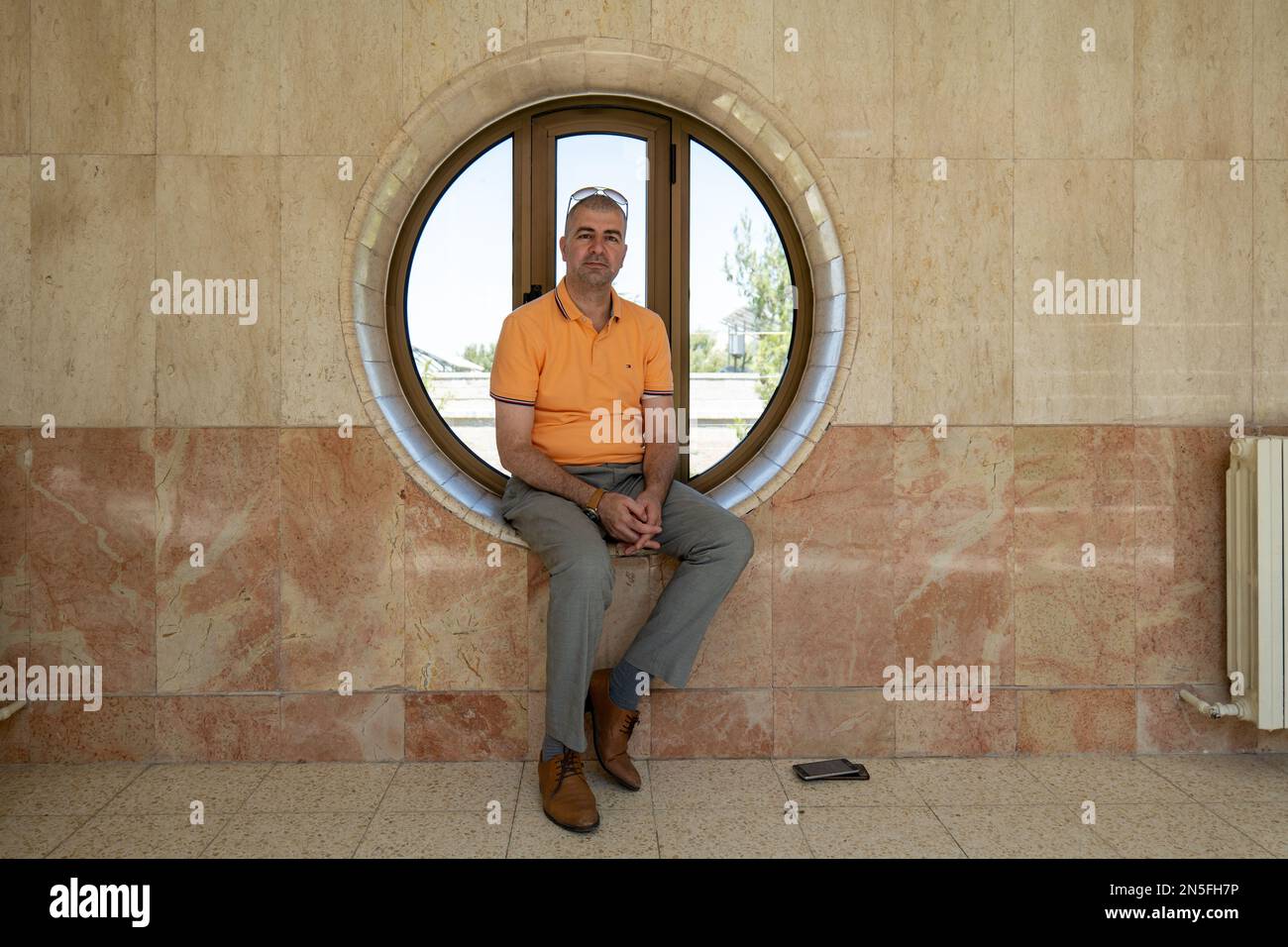 Bethlehem, Westjordanland, Palästina - 22. Juli 2022: Arabischer Mann in orangefarbenem Hemd und grauer Hose sitzt in einem runden Fenster in einer Steinmauer Stockfoto