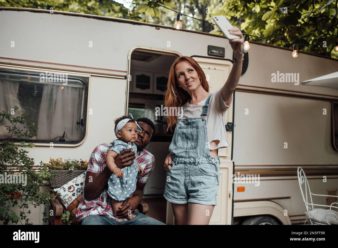 Gemischte Familien mit dunkel gehäuteter Kleinkindertochter vor dem Wohnwagen machen Fotos für soziale Medien. Ein afroamerikanischer Mann, seine Frau und sein kleines Kind Stockfoto