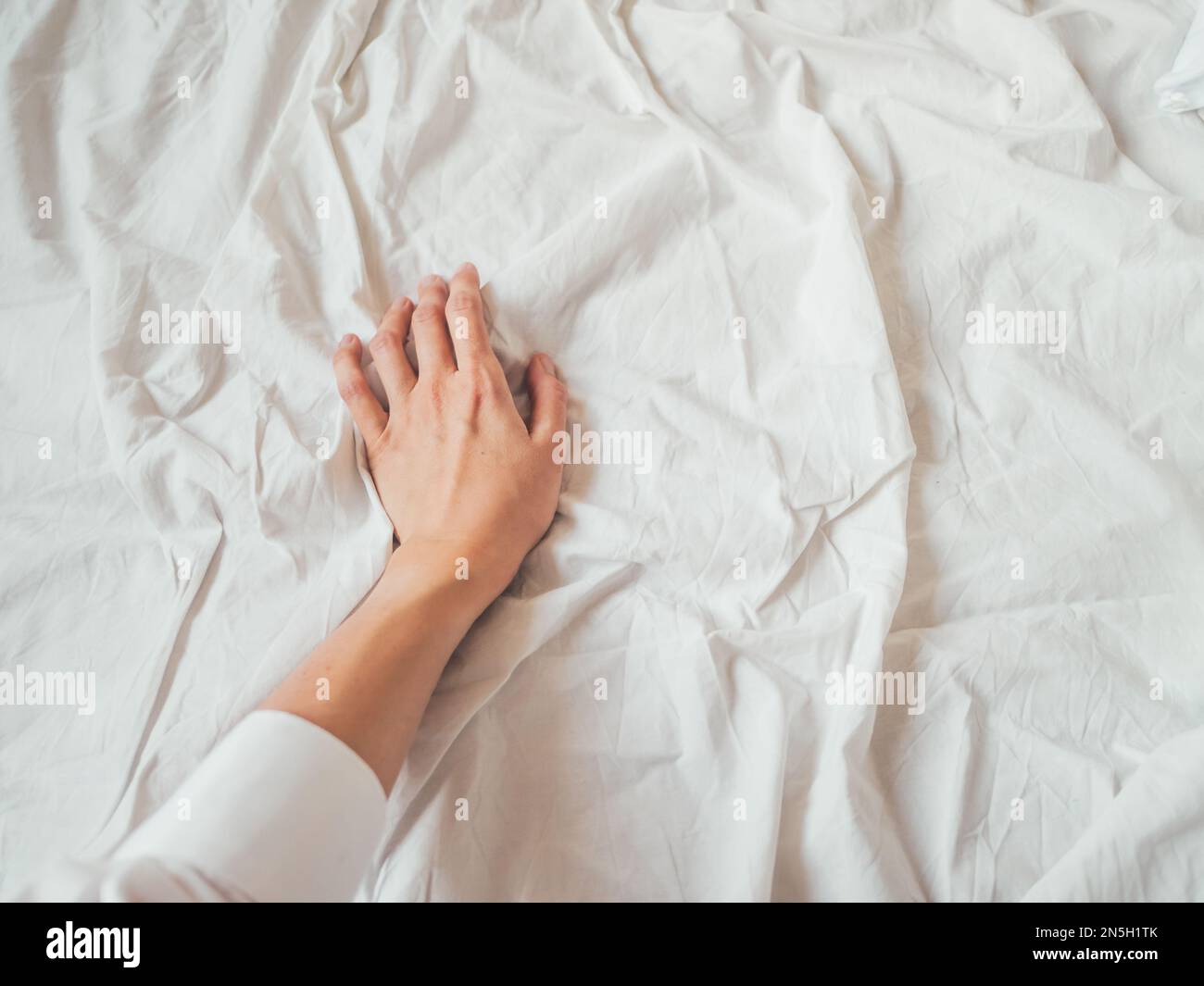 Die Hand der Frau auf dem Hintergrund des zerknitterte Bettlaken. Gefittertes weißes Leinen. Die Frau berührt das ungemachte Bett. Gemütliches, unperfektes Haus. Nicht ideal gemütliches Zuhause. Stockfoto