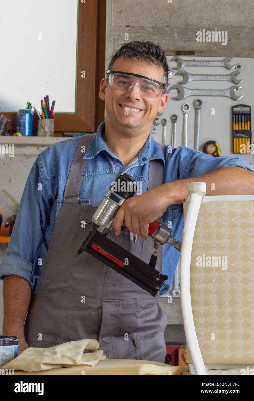 Lächelnder Polsterer in seiner Werkstatt mit verschiedenen Werkzeugen und Nagelpistole nach Restaurierung eines alten Stuhls. Stockfoto