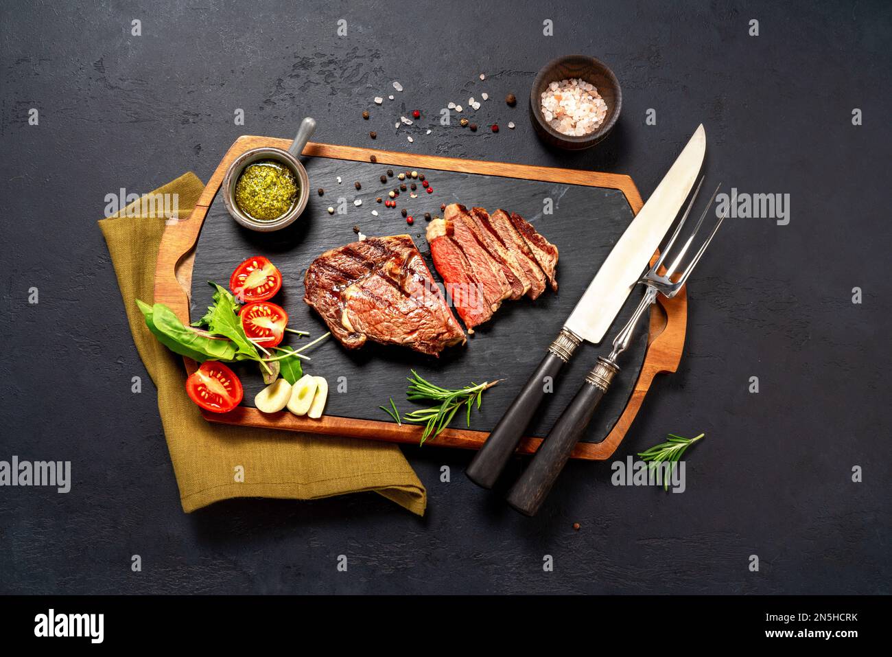 Gegrilltes Ribeye-Steak, Chimichurri-Sauce, Kräuter und Gewürze auf einem dunklen Tisch. Steak in Scheiben auf dunklem Brett für Steak Stockfoto