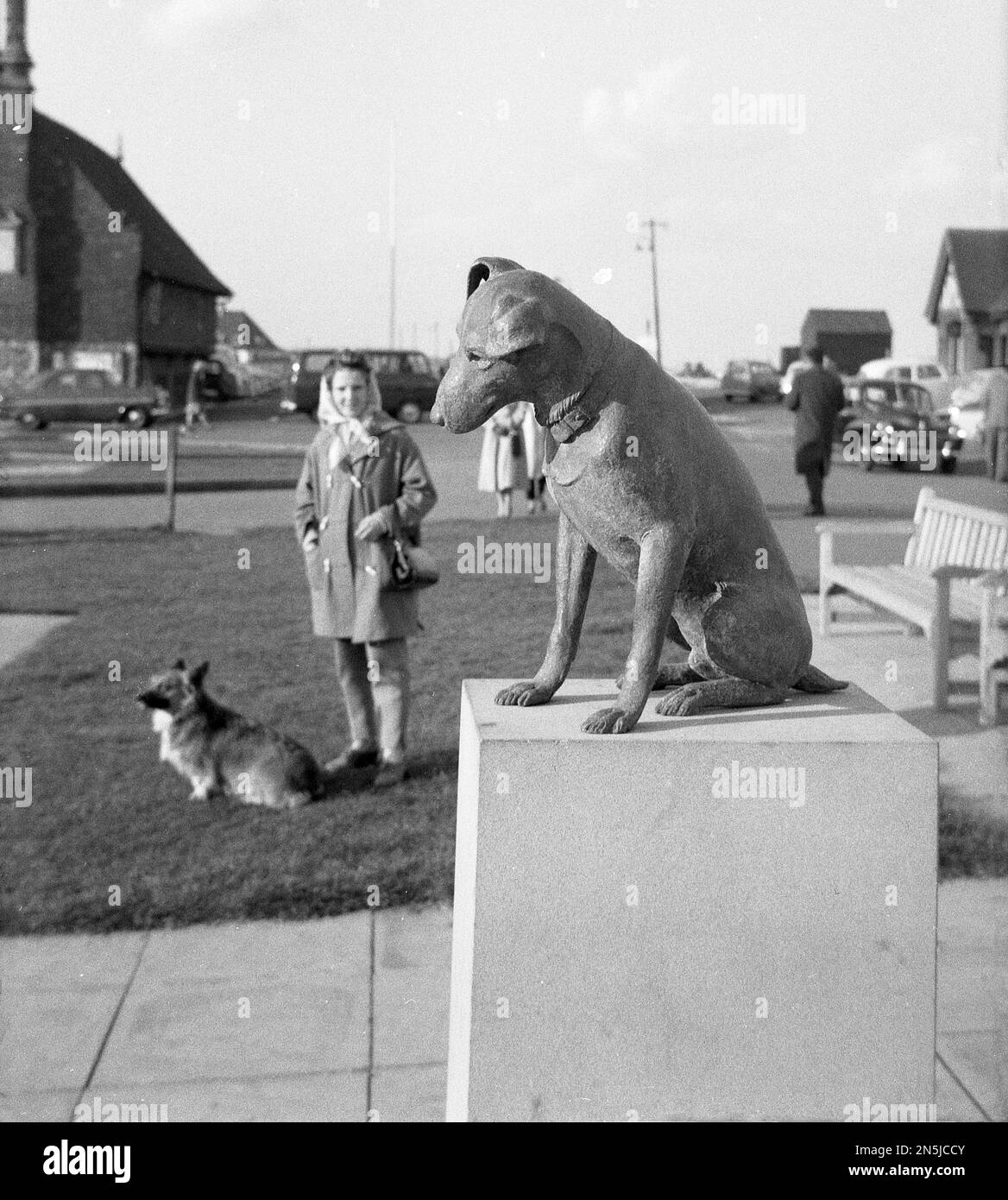 1963, historisch, eine Dame mit ihrem kleinen Hund neben ihr, stehend, auf eine Statue eines Hundes, 'Snooks' auf dem Marktplatz, Aldeburgh, Suffolk, England, Großbritannien. Die Bronzestatue wurde 1961 zum Gedenken an den örtlichen Aldeburgh GP Dr. Robin Acheson errichtet, als sein Hund Snooks, der dem Arzt folgte, während er seine Anrufe machte, zu einem vertrauten Anblick rund um die Küstenstadt wurde. Stockfoto