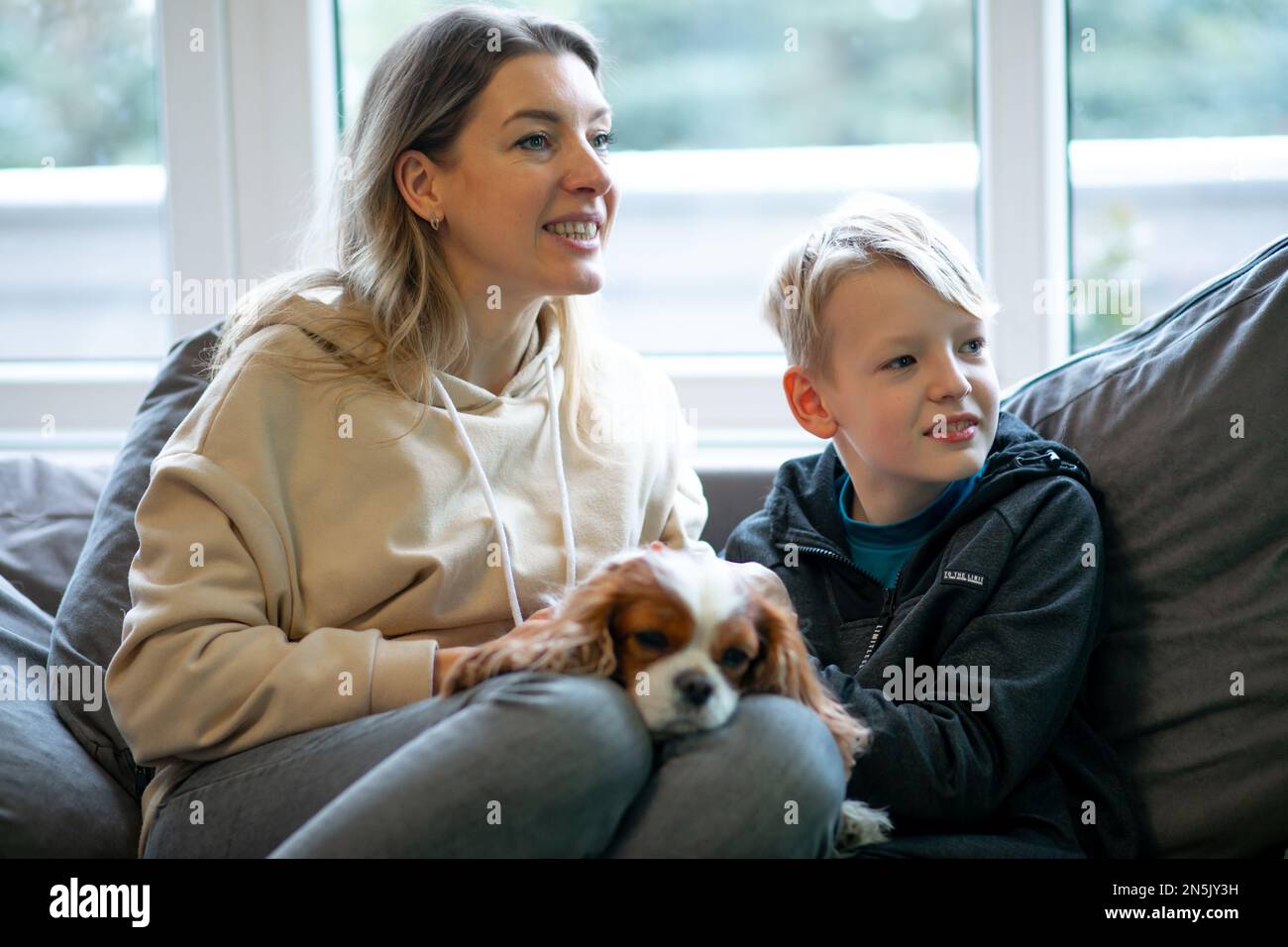 Mutter und Sohn sitzen auf dem Sofa neben dem Fenster mit Spaniel. Familienporträt in einem gemütlichen Zuhause. Trauriger Welpe, der auf weiblichen Beinen liegt. Stockfoto