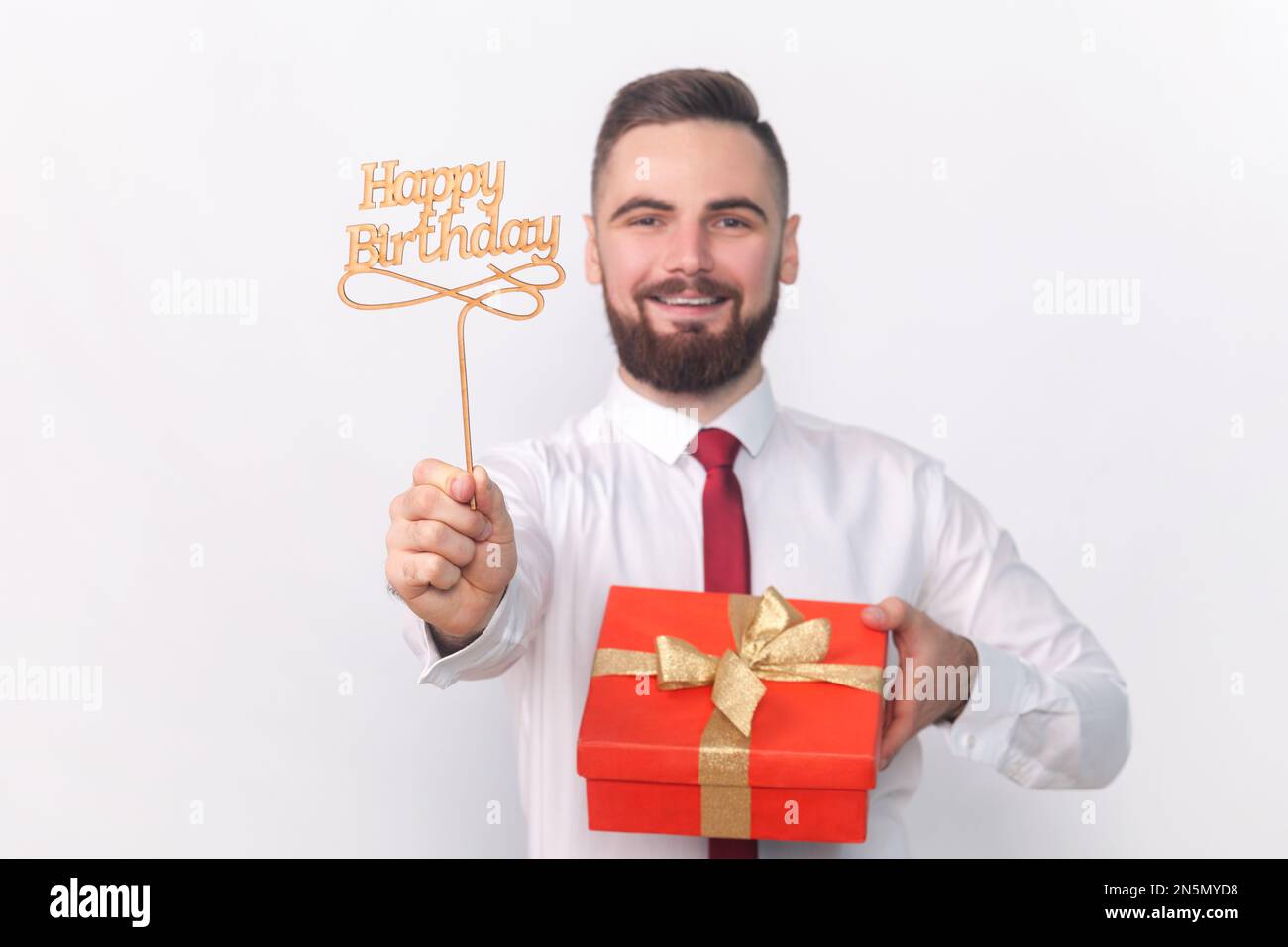 Portrait eines lächelnden, zufriedenen, gutaussehenden Mannes in weißem Hemd, der Partyauflagen hält, mit Happy Birthday-Inschrift und roter Geschenkbox. Studioaufnahmen im Innenbereich isoliert auf grauem Hintergrund. Stockfoto