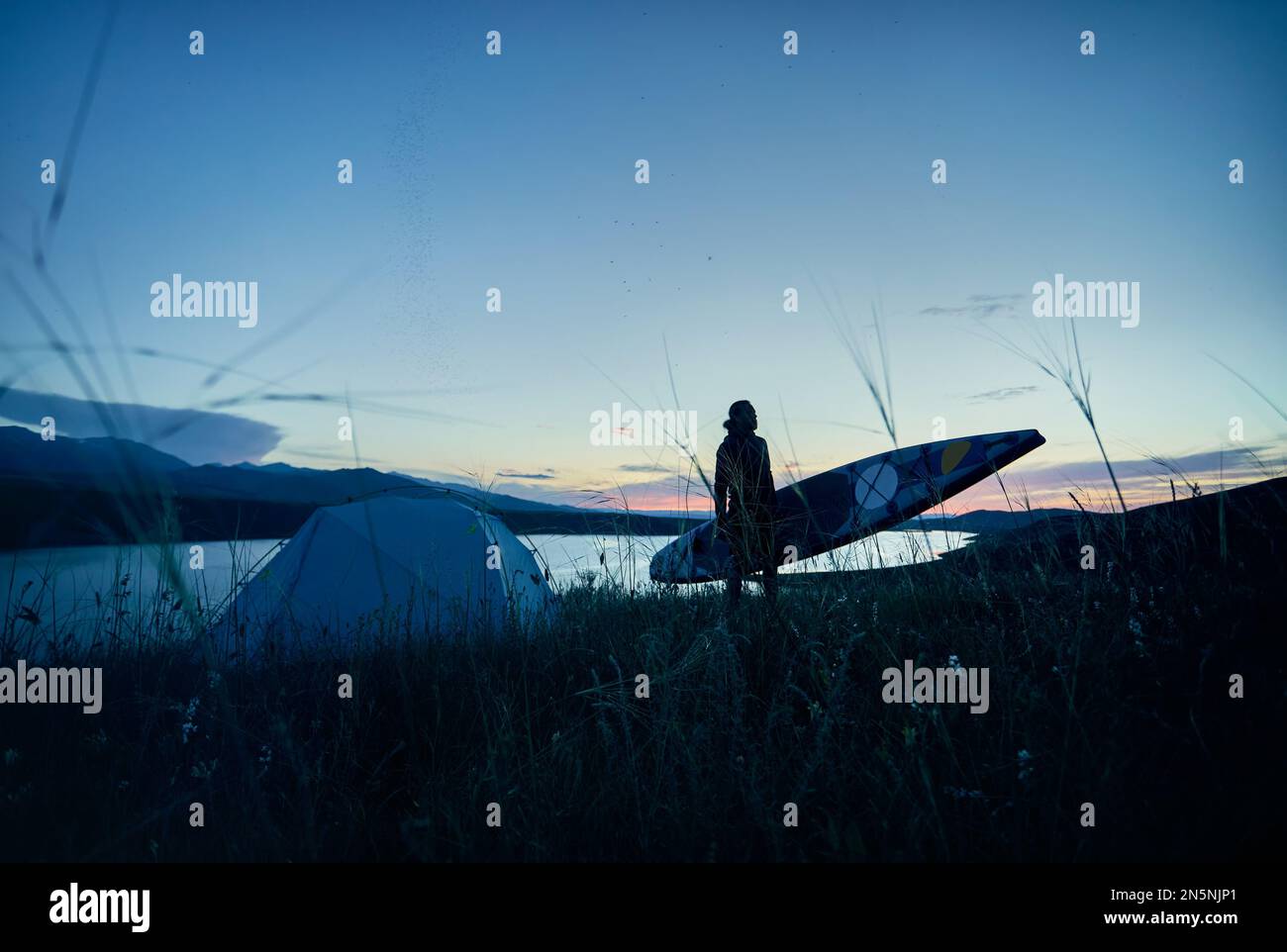 Mann in Silhouette mit Paddel und SUP-Brett in der Nähe des Zelts, Camping am See bei Sonnenuntergang in Kasachstan. Stehpaddelboarden aktive Outdoor-Erholung in n Stockfoto