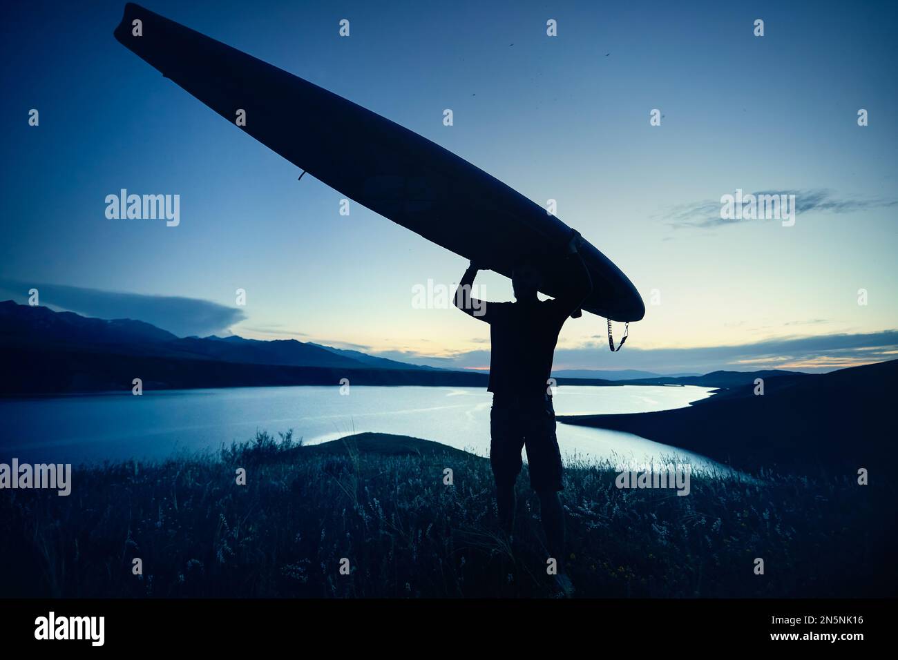 Ein Mann in Silhouette, der bei Sonnenuntergang in Kasachstan ein SUP-Brett auf dem Kopf hält. Stehpaddeln Outdoor-Aktivitäten in der Natur. Stockfoto