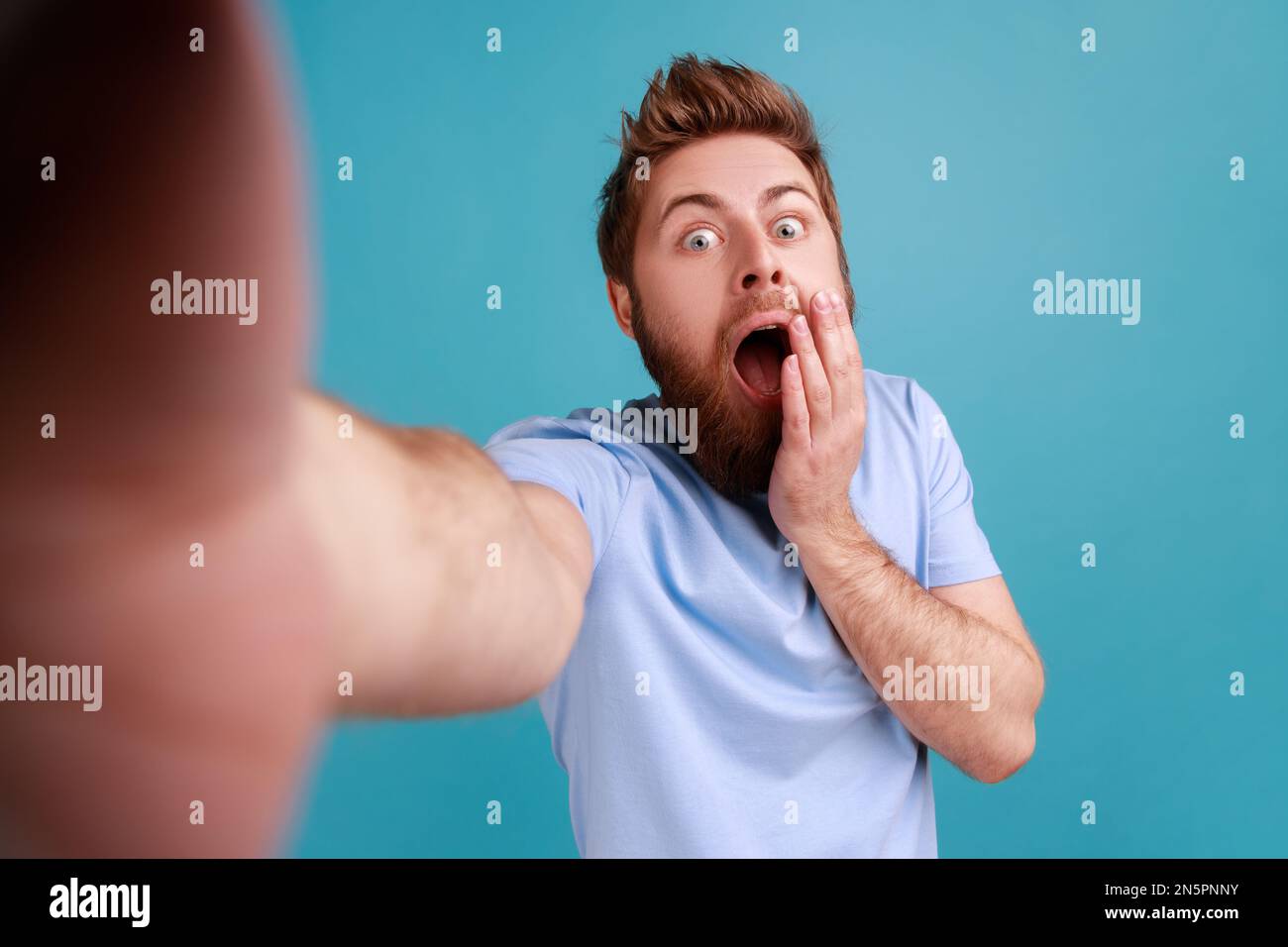 Porträt eines bärtigen Mannes, der Selfie macht oder Videoanrufe macht, mit schockiertem Gesichtsausdruck in die Kamera schaut, Blickwinkel des Fotos. Innenstudio, isoliert auf blauem Hintergrund. Stockfoto