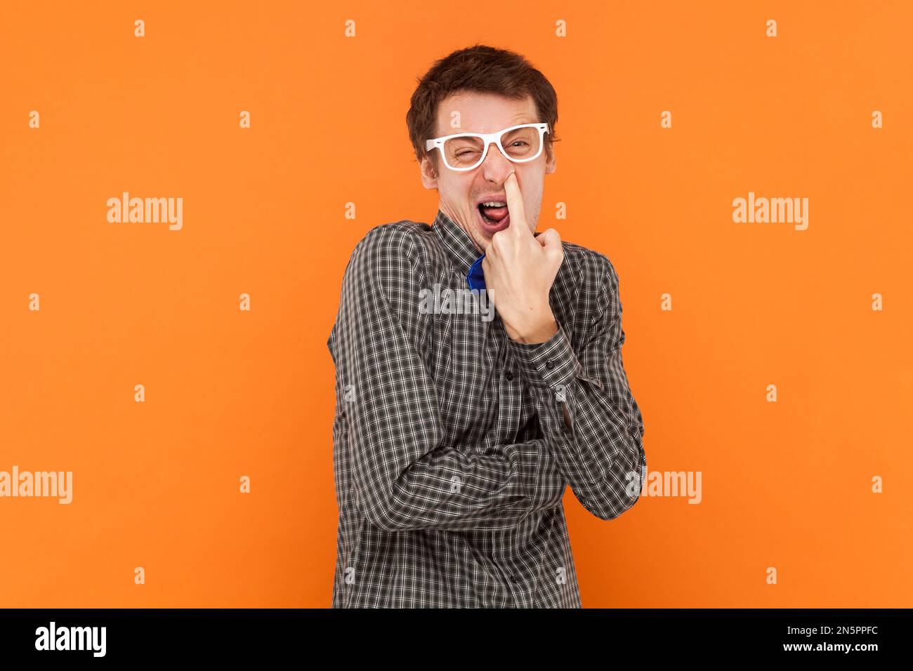 Porträt eines Mannes, der sich den Finger in die Nase steckt, herumalbert, schlechte Angewohnheiten, respektloses Verhalten, trägt ein Hemd mit blauer Fliege und einer weißen Brille. Innenstudio, isoliert auf orangefarbenem Hintergrund Stockfoto