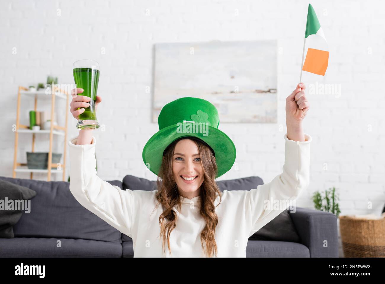Fröhliche junge Frau mit grünem Hut und irischer Flagge und Bier zu Hause Stockfoto