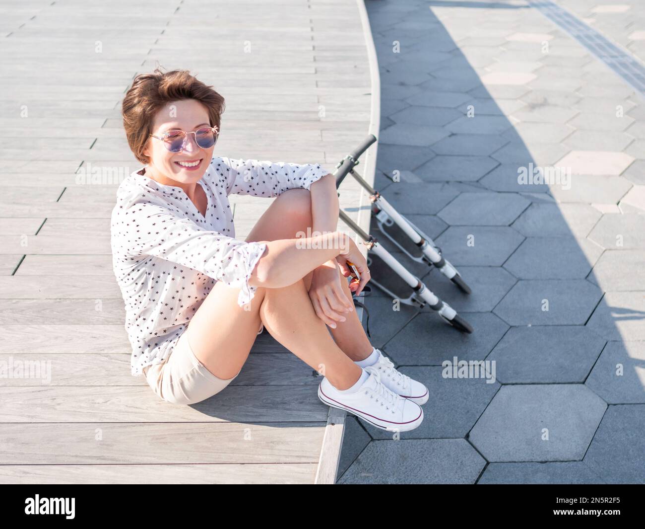 Der Wind rührt kurzes Haar einer Sommersprossen in einer farbenfrohen Sonnenbrille. Lächelnde Frau in einer offenen Holzszene. Sommerliche Stimmung. Aufrichtige Emotionen. Kick-Roller Stockfoto