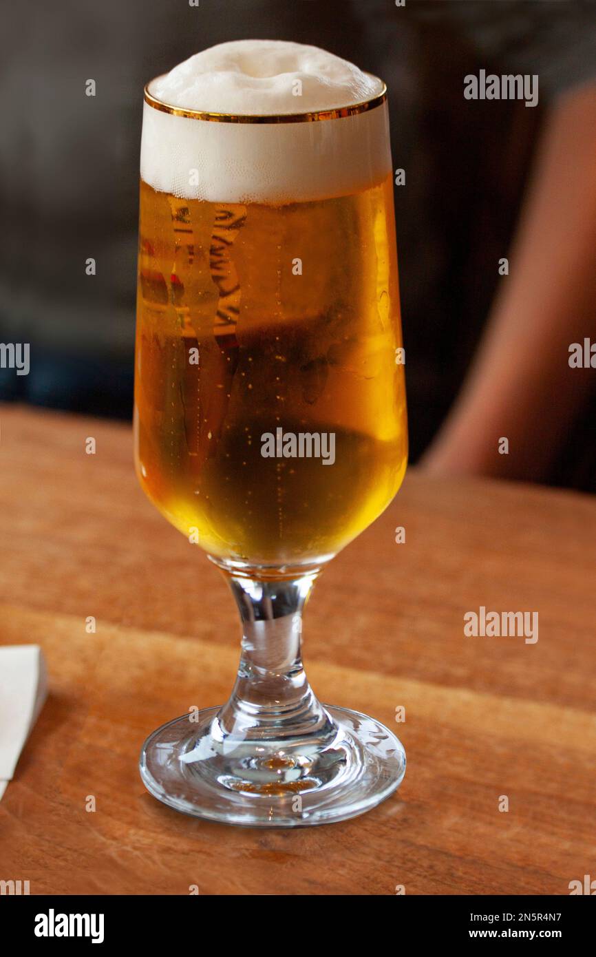 Gekühltes Glas Bier mit Kondensationstropfen auf Glas auf einem Holztisch. Atmosphärisches Bild mit verschwommener Person im Hintergrund. Stockfoto