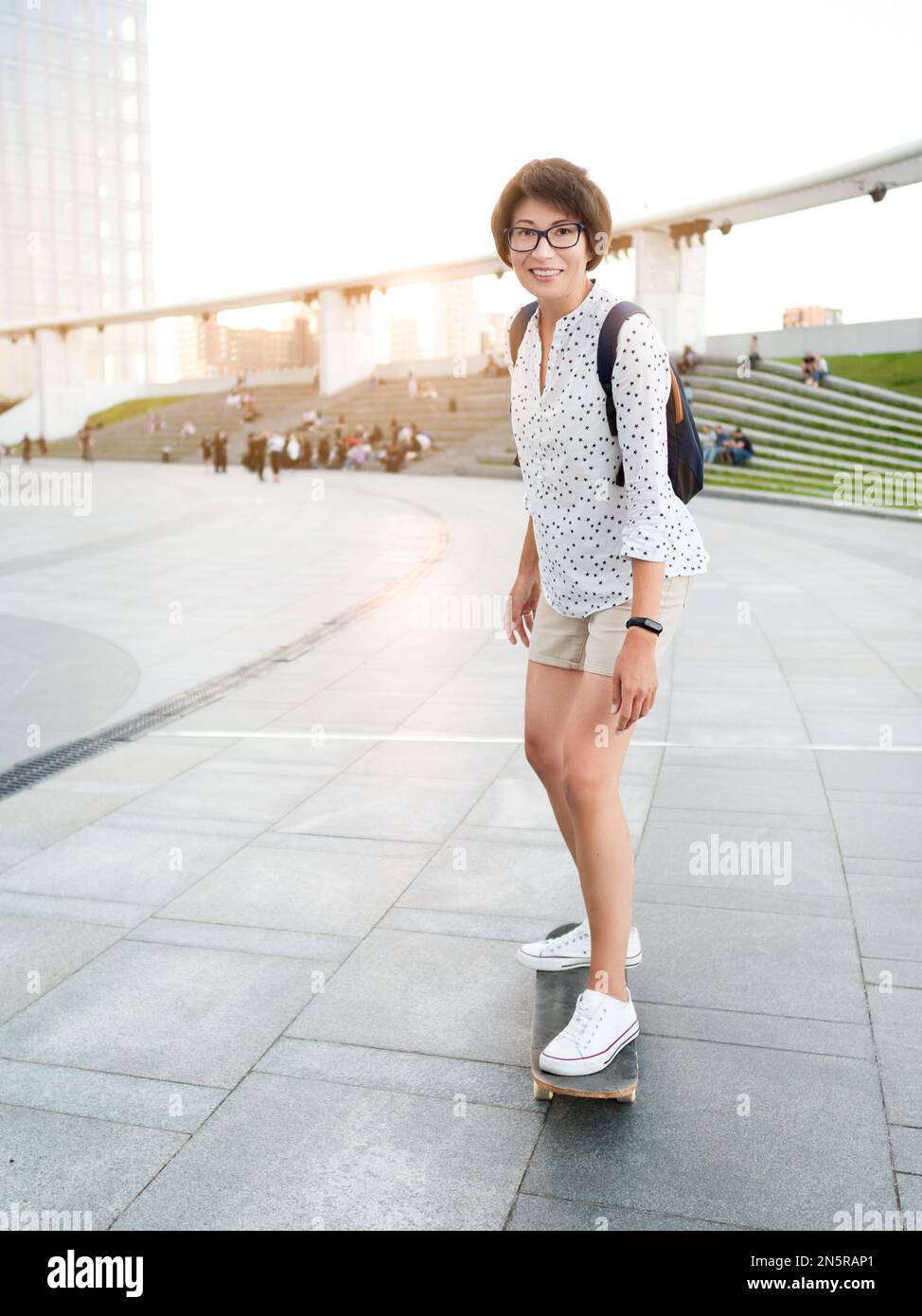 Frau fährt Skateboard auf dem Stadtplatz bei Sonnenuntergang. Sommerliche Stimmung. Neue Dinge im Erwachsenenalter lernen. Millennial lernt Skaten. Gesunder Lebensstil. Stockfoto
