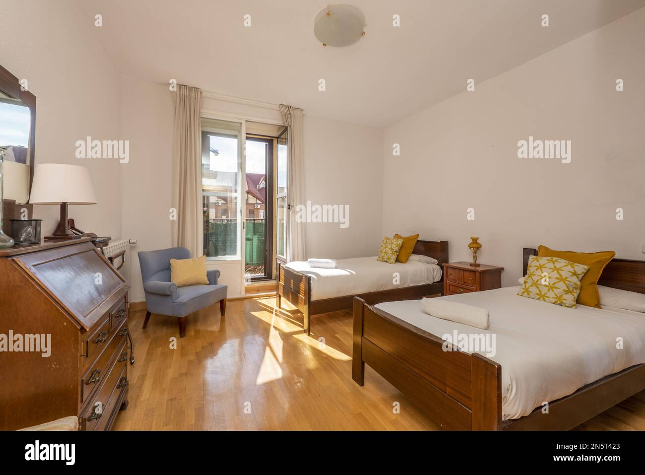 Schlafzimmer mit zwei Holzeinzelbetten, Schreibtisch mit Schubladen und geschlossenem Klappdach, blauem Sessel und Balkon mit Aussicht Stockfoto