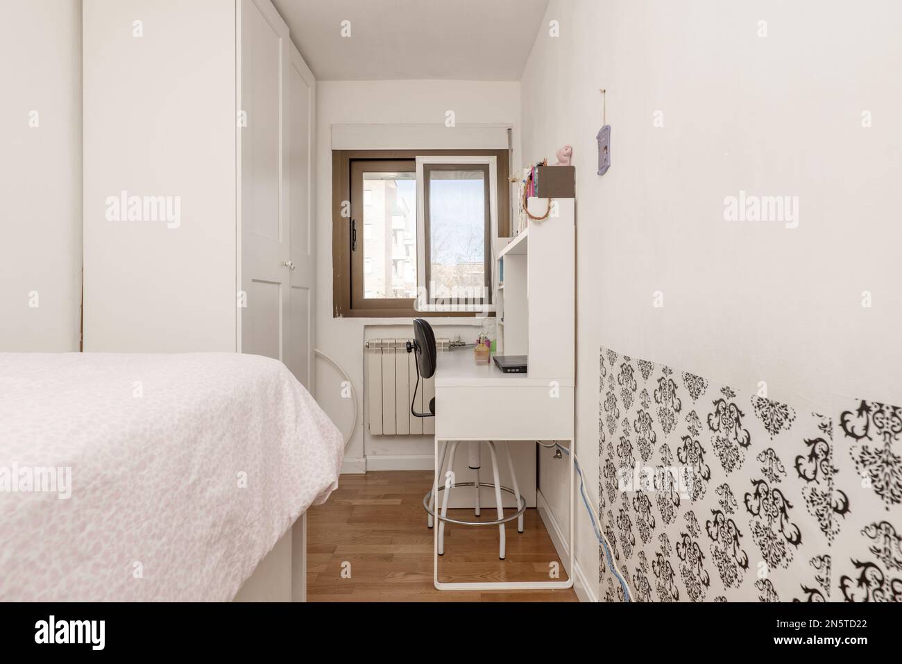 Schmales Schlafzimmer mit einem Einzelbett, einem weißen Schreibtisch, einem Kleiderschrank mit weißen Türen und einem Aluminiumfenster Stockfoto