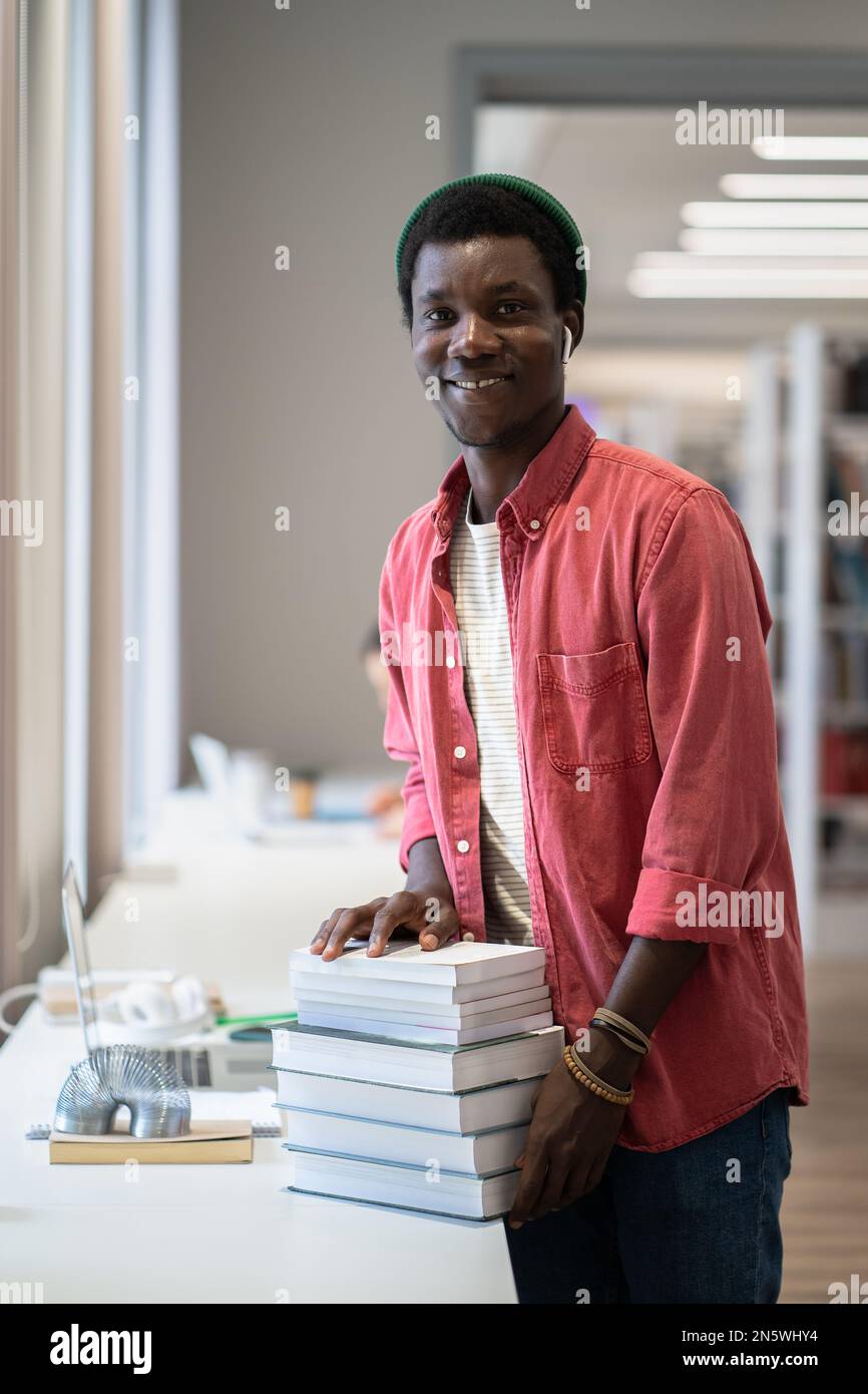 Junger, fröhlicher afroamerikanischer Bibliothekar, der mit einem Stapel Bücher in der öffentlichen Bibliothek steht Stockfoto
