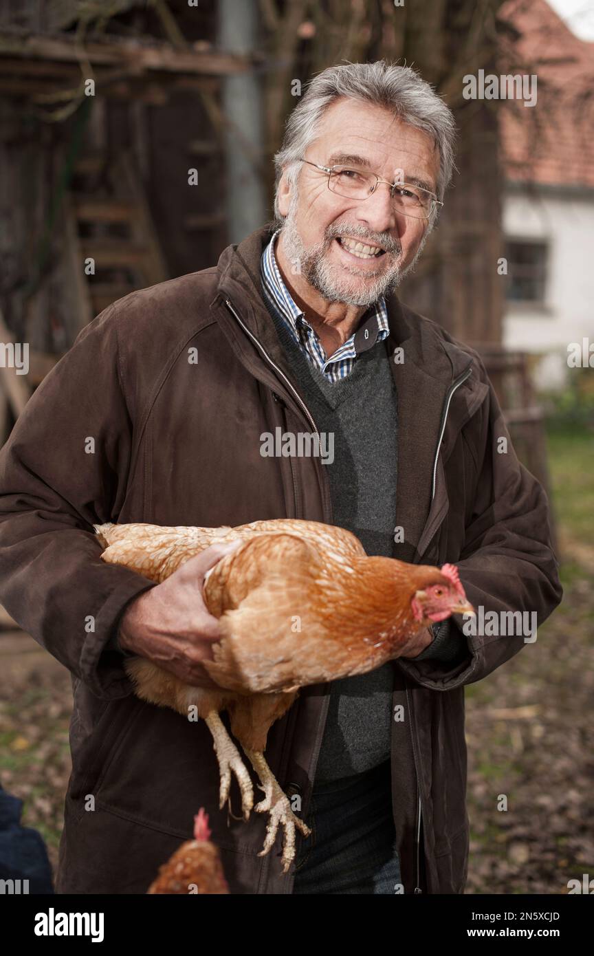 Porträt eines Seniorenmannes, der Hühnervögel auf dem Bauernhof hält und lächelt, Bayern Stockfoto
