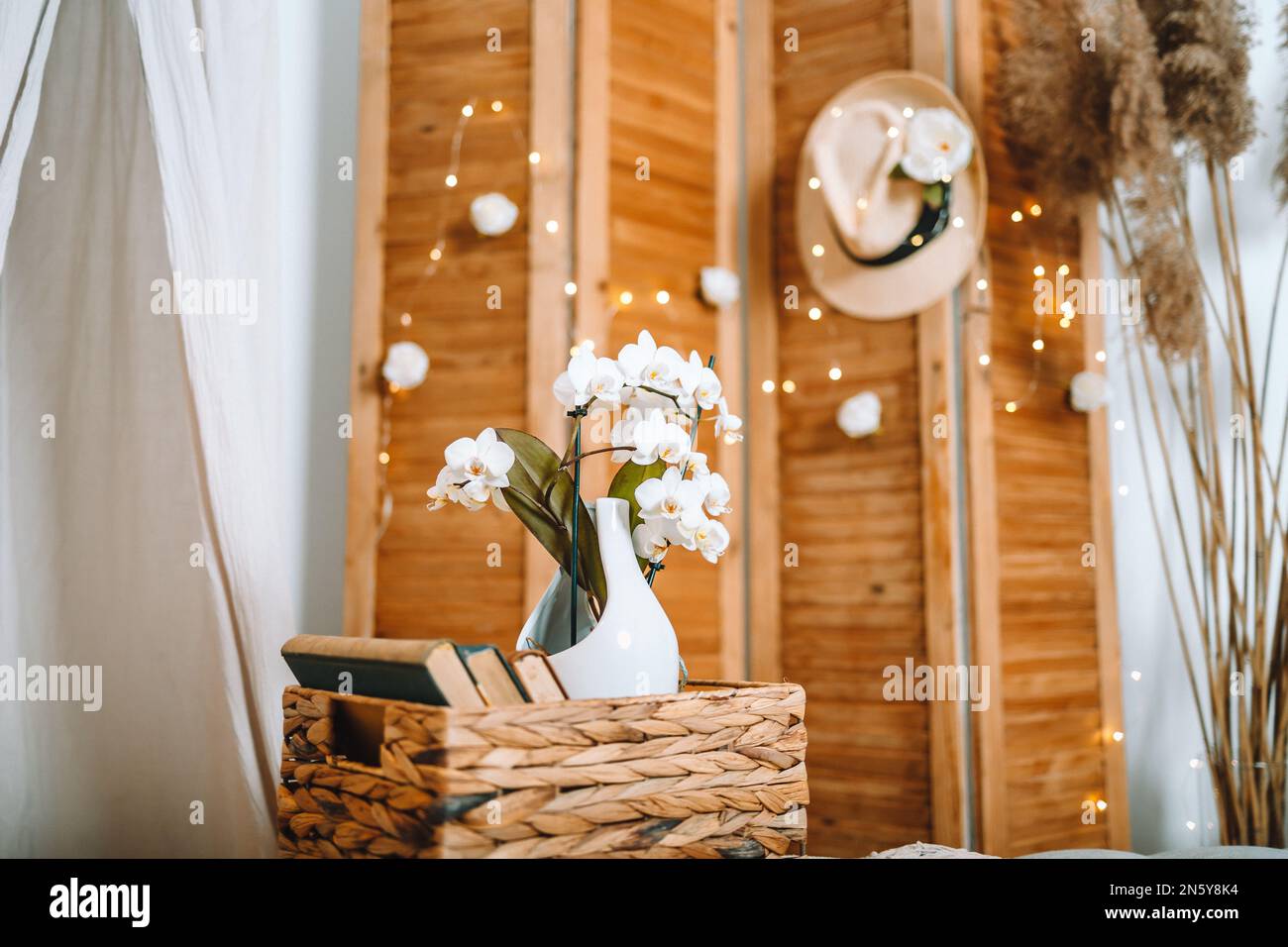 Weißes Orchideen in Weiß und Beige dekoriertes gemütliches Zimmer, beigefarbener Hut auf einem natürlichen Klappbildschirm mit weißen Rosen, Pampas und warmen LED-Lampen, gemütlich und weich Stockfoto