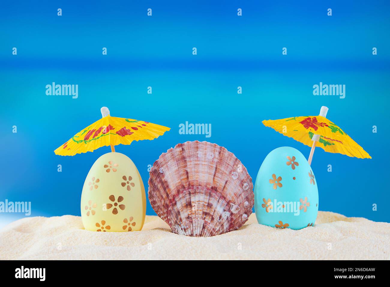Frohe Ostern. Gelbe und blaue Eier mit Muschelschale unter Sonnenschirmen am Sandstrand des Meeres oder Ozeans. Die Feiertagspostkarte in heißem Land. Reisen, Sprotte Stockfoto