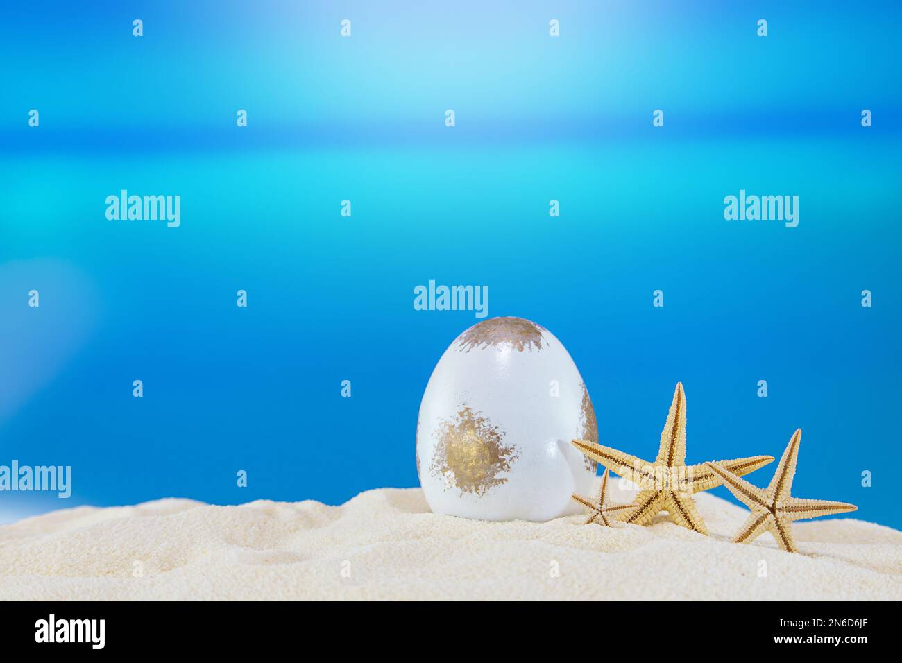 Weißes goldenes Ei mit Seesternen am Sandstrand des Meeres oder Ozeans. Urlaubspostkarte im sonnigen Land. Frohe Ostern. Reisen, Frühling, Sommer. Speicherplatz kopieren Stockfoto