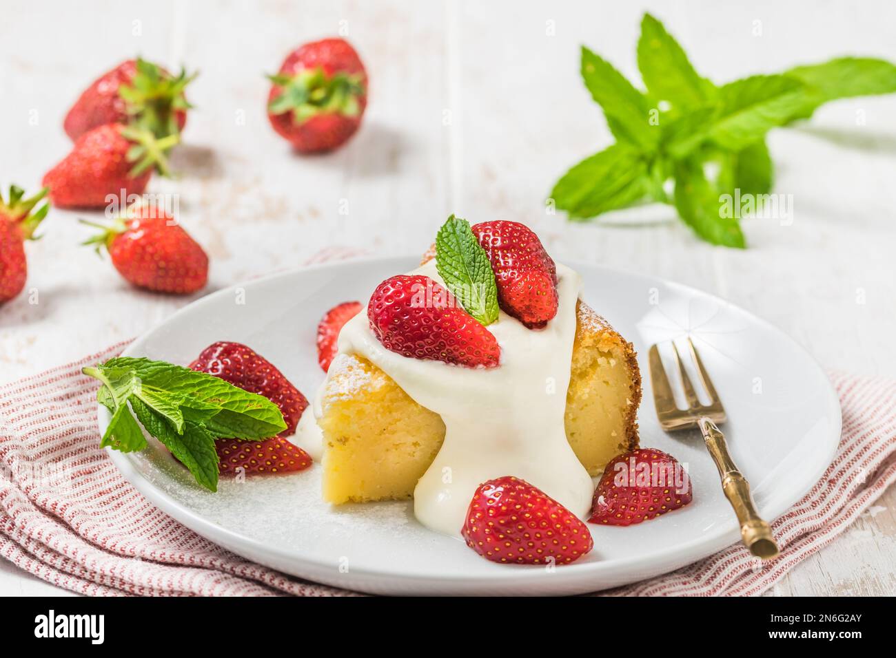 Ein Stück hausgemachter Käsekuchen mit Schlagsahne und frischen Erdbeeren auf weißem Holzhintergrund, dekoriert mit frischen Minzblättern Stockfoto