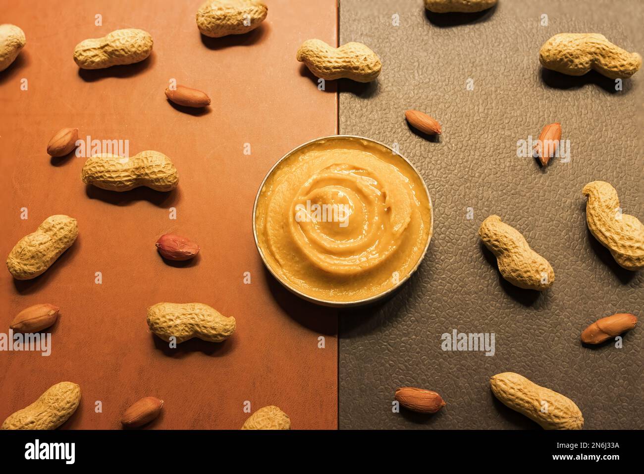 Kopfaufnahme eines Bechers Erdnussbutter auf einem zweifarbigen Hintergrund mit zufällig angeordneten ganzen geschälten Erdnüssen Stockfoto
