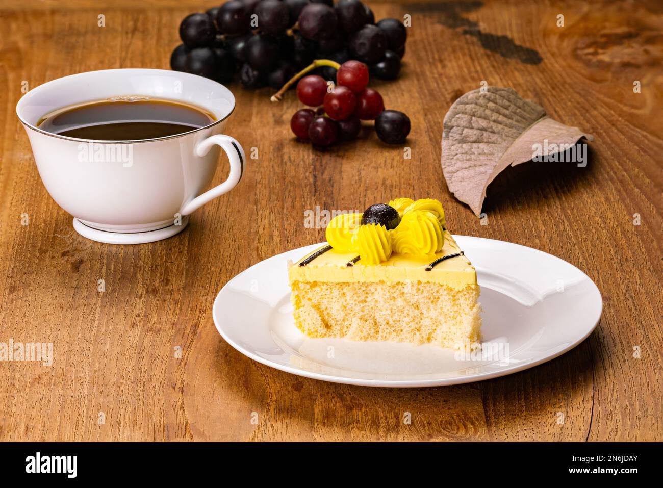 Frühstück mit hausgemachtem, köstlichem Kuchen mit gelber Blumencreme in weißer Keramikschale mit einer Tasse schwarzem Kaffee und Bündeln reifem Gummi Stockfoto