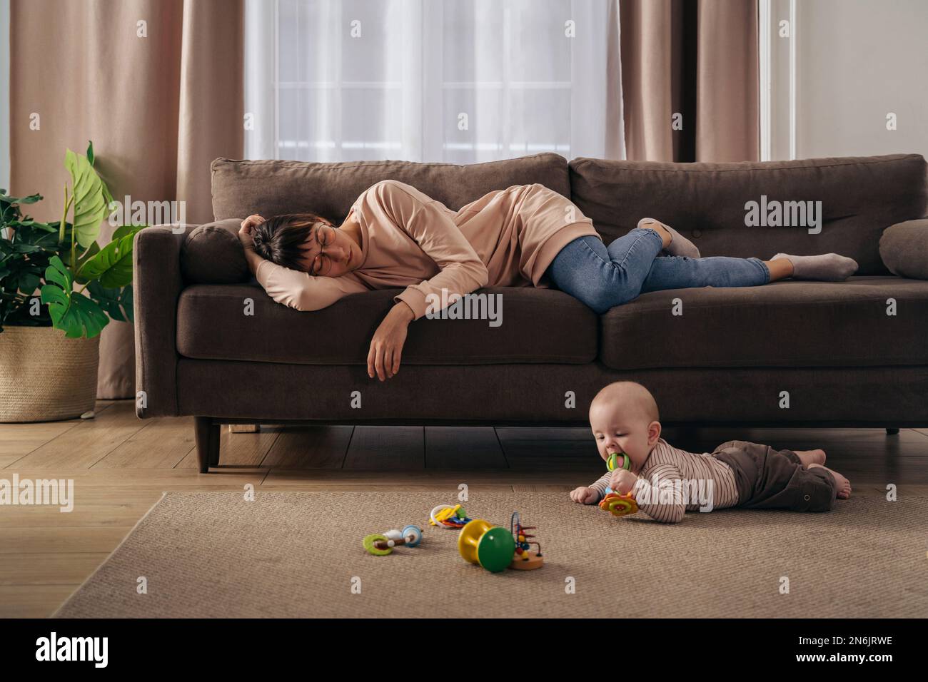 Junge, müde Mutter, die unter Schlafmangel leidet, schläft auf dem Sofa, während ihr kleines Baby auf dem Boden spielt. erschöpfte mutter mit postnataler Depression, will nicht mit ihrem Sohn spielen Stockfoto