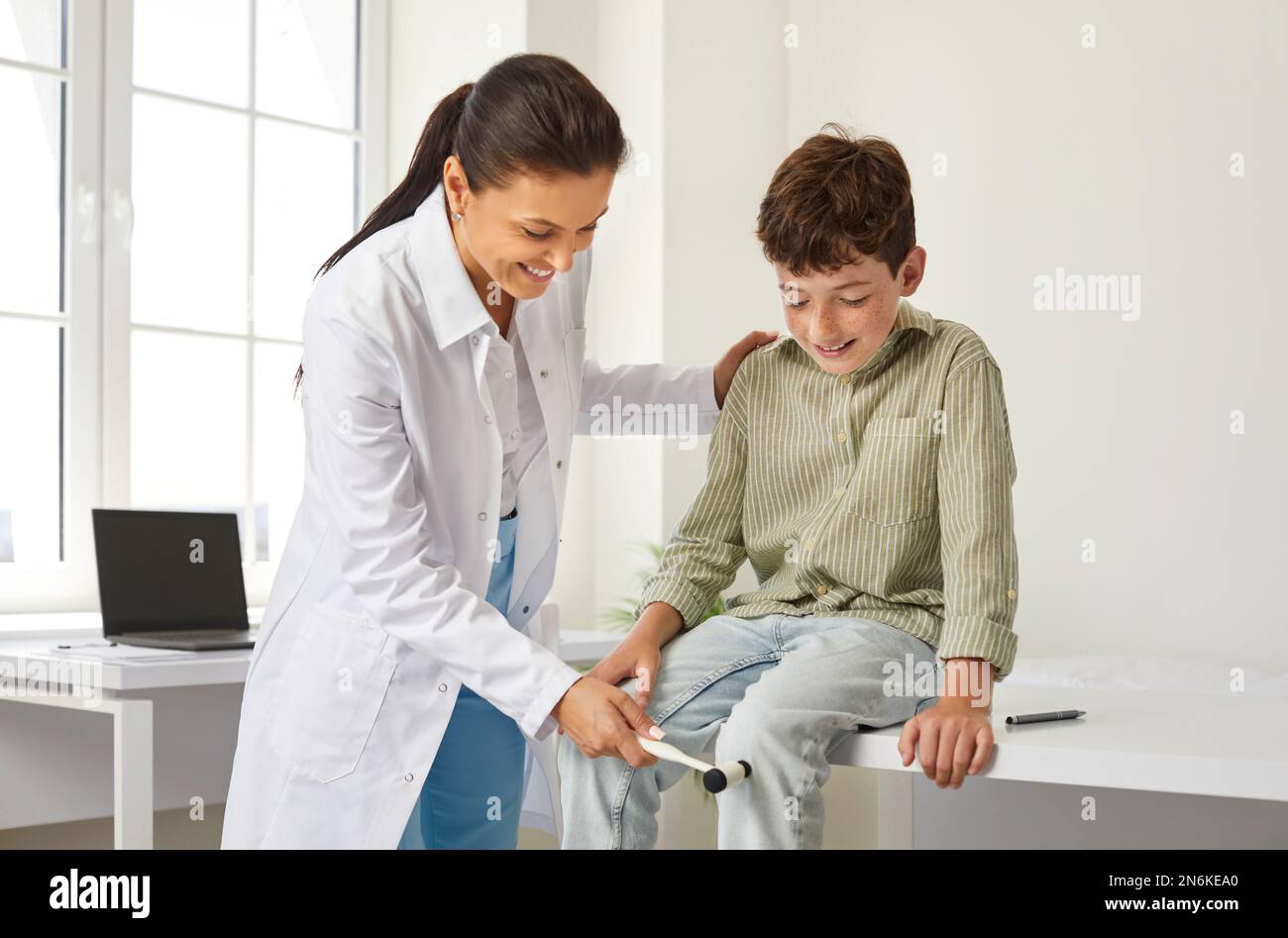 Doktor untersucht ein glückliches kleines Kind und benutzt einen Hammer, um seinen Knieflex zu überprüfen Stockfoto