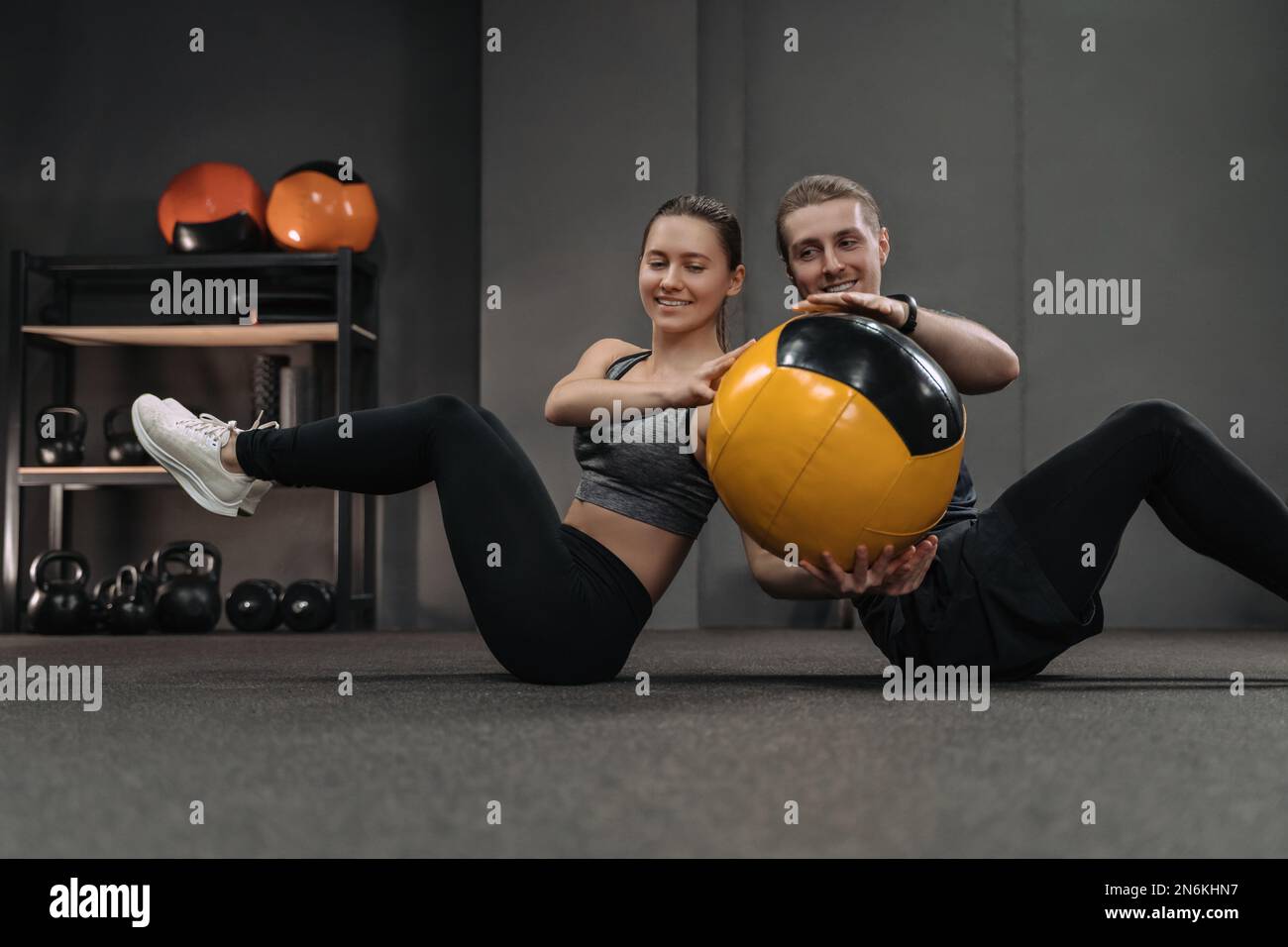 Gesundes junges Paar, das bauchmuskelübungen mit Medizinball im Dark Gym macht. Das Paar lehnt sich Rücken an Rücken auf dem Boden an und trainiert, indem es den Ball an den anderen weitergibt. Sportliche Frauen und Männer trainieren zusammen Stockfoto