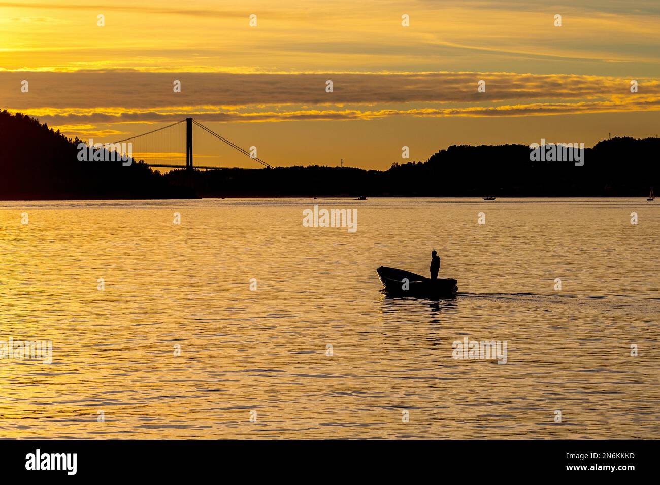 Ein Mann in einem kleinen Boot, in Byfjorden, bei Sonnenuntergang. Außerhalb des Hafens Bergen, Norwegen. Askøy-Brücke im Hintergrund Stockfoto
