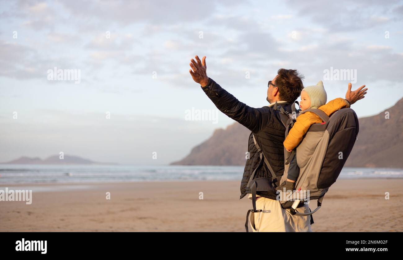 Der Vater erhebt sich die Hände zum Himmel und genießt dabei die reine Natur, während er seinen kleinen Sohn im Rucksack am windigen Sandstrand von Famara, Lanzarote Islan, trägt Stockfoto