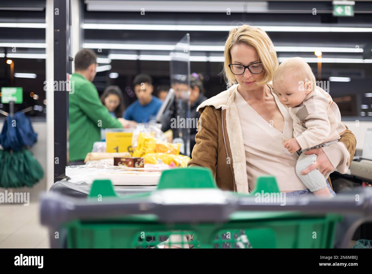 Mutter kauft mit ihrem kleinen Jungen ein und hält das Kind, während sie Produkte an der Kasse im Supermarkt stapelt Stockfoto
