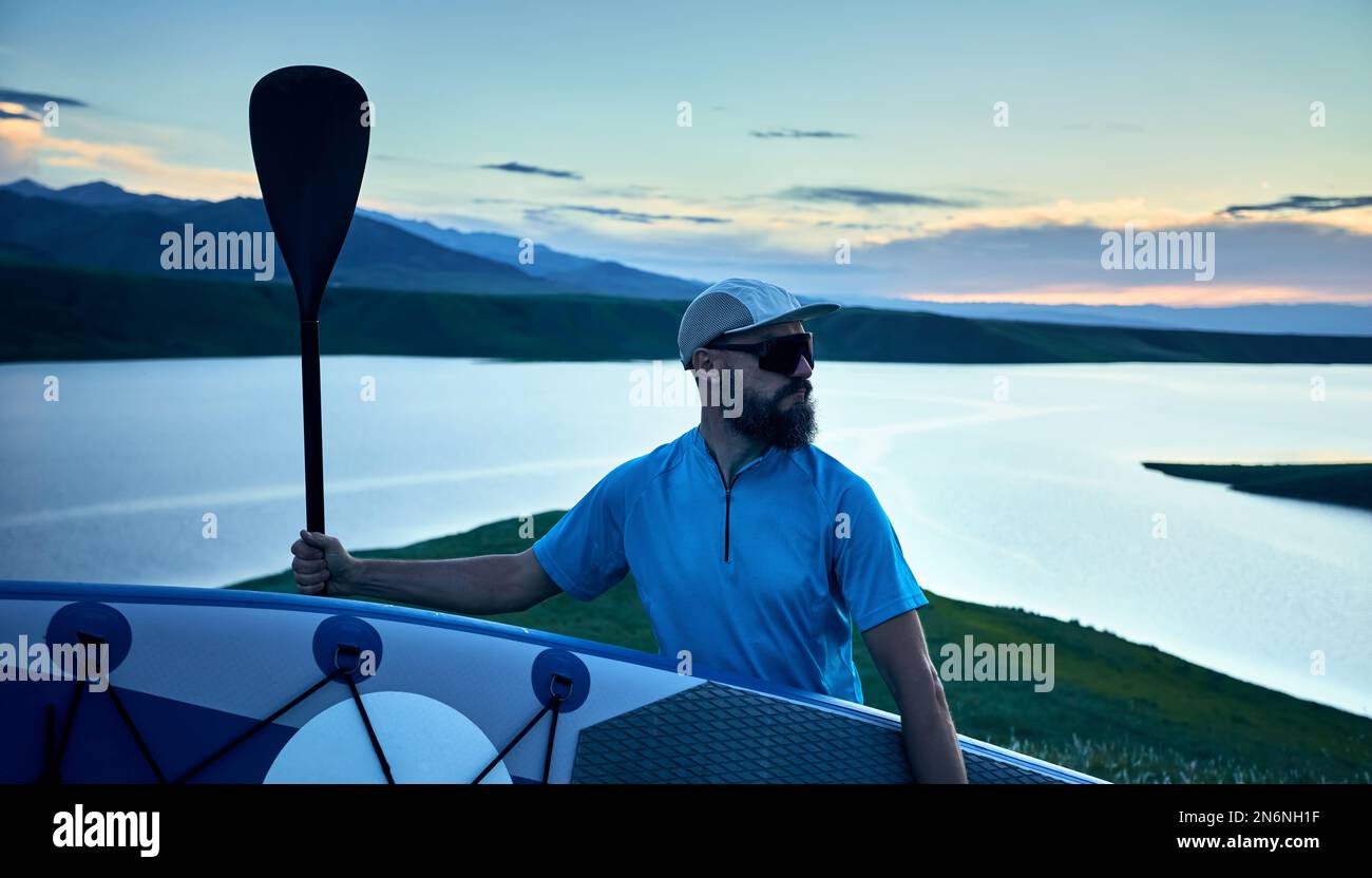 Porträt eines bärtigen Mannes in Silhouette mit SUP-Paddel in der Nähe des Sees bei Sonnenuntergang in Kasachstan. Stand Up Paddle Boarding Outdoor aktiv Erholung i Stockfoto