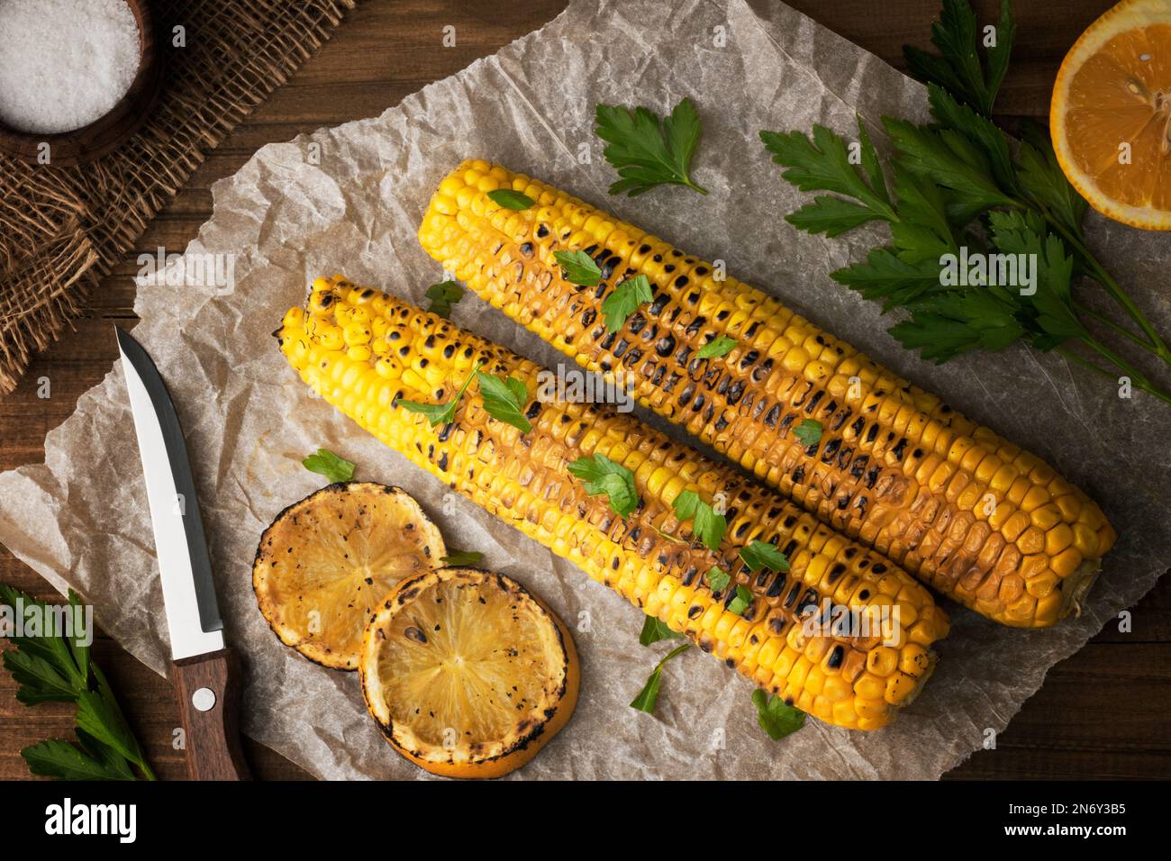 Köstlicher gegrillter mexikanischer Mais mit Petersilie, gegrillter Zitrone und Salz. Blick von oben auf den Küchentisch mit gegrilltem Zuckermaiskolben auf Backpapier. Veganes Restaurant Stockfoto