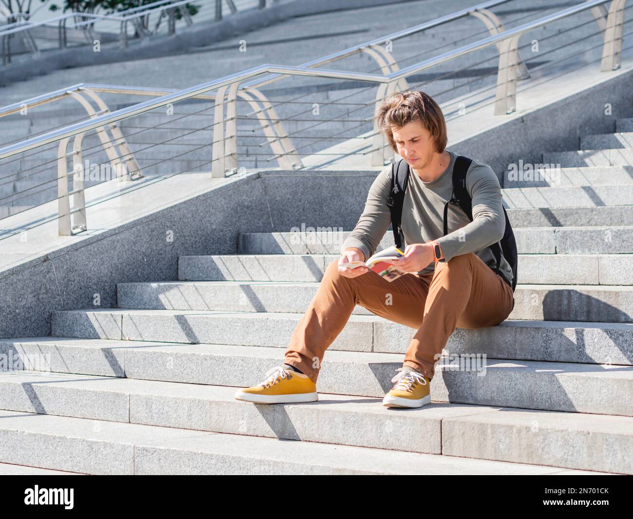 Der junge Mann sitzt auf einer Steintreppe im Park und liest den Reiseführer. Alleinreisen durch die Stadt. Stadttourismus. Moderne architektonische Wahrzeichen. Stockfoto