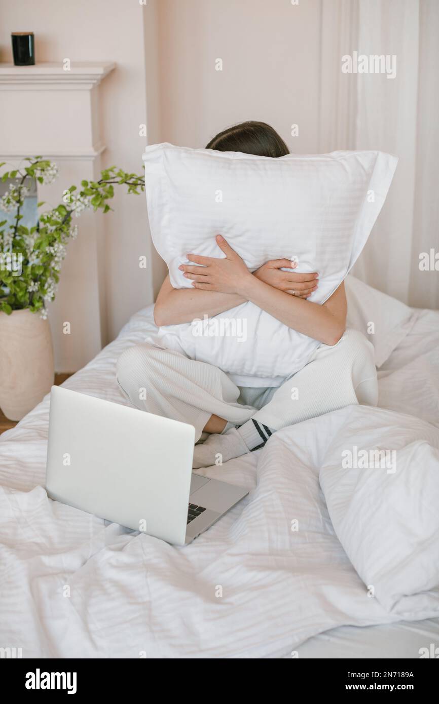 Frau, die im Bett vor ihrem Laptop sitzt und ein Kissen kuschelt Stockfoto