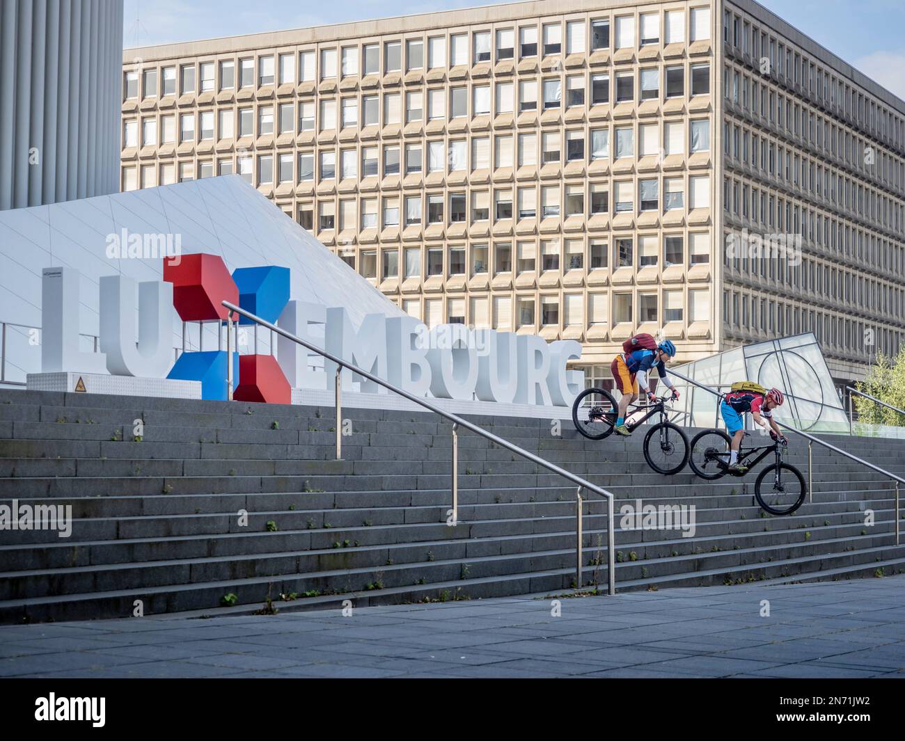 E-Mountainbiker auf den Stufen der Philharmonie mit Schriftzug Luxemburg. Im Hintergrund das Schumann-Gebäude, das in den 70s Jahren erbaut wurde, um die ersten EU-Institutionen in Luxemburg zu beherbergen. Stockfoto