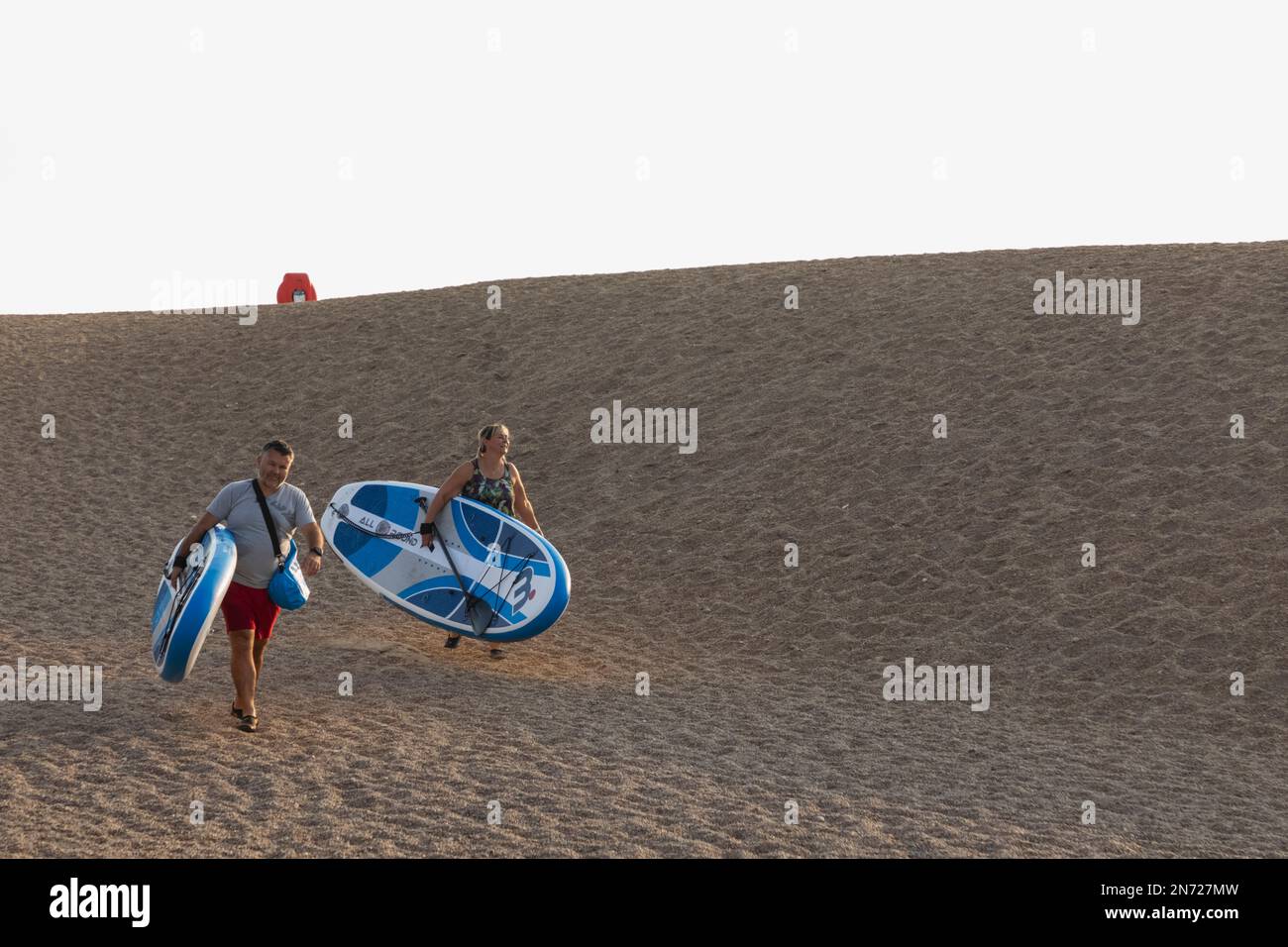 England, Dorset, Bridport, West Bay, ein Paar mittleren Alters, das Paddle Boards am Strand trägt Stockfoto