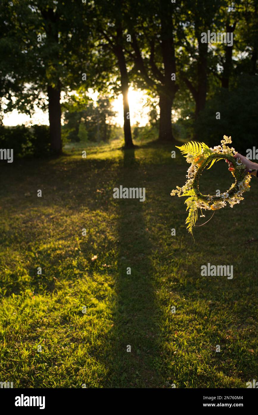 Hand mit einer Blumenkrone während einer Sommerfeier in Lettland. Die Abendsonne scheint durch die Bäume im Hintergrund. Stockfoto