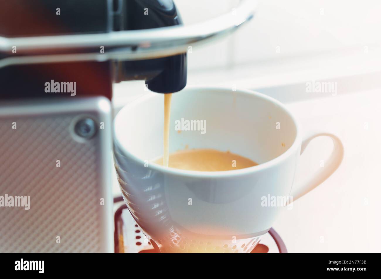 Frischer heißer Kaffee fließt aus einer modernen Kapselmaschine in eine weiße Porzellantasse, farbige Lichtreflexe, heller Hintergrund mit ausgewähltem Kopierbereich Stockfoto