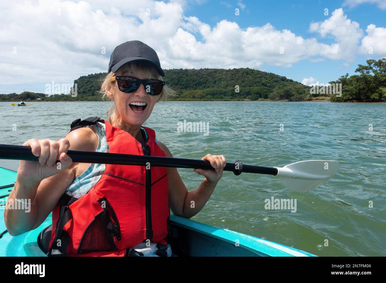 Eine Dame in ihren 60s Jahren saß in einem Kanu und hielt ein Paddel, lächelte und lachte und sah sehr glücklich auf einem Fluss aus. Sie trägt eine Sonnenbrille und eine Baseballmütze. Stockfoto