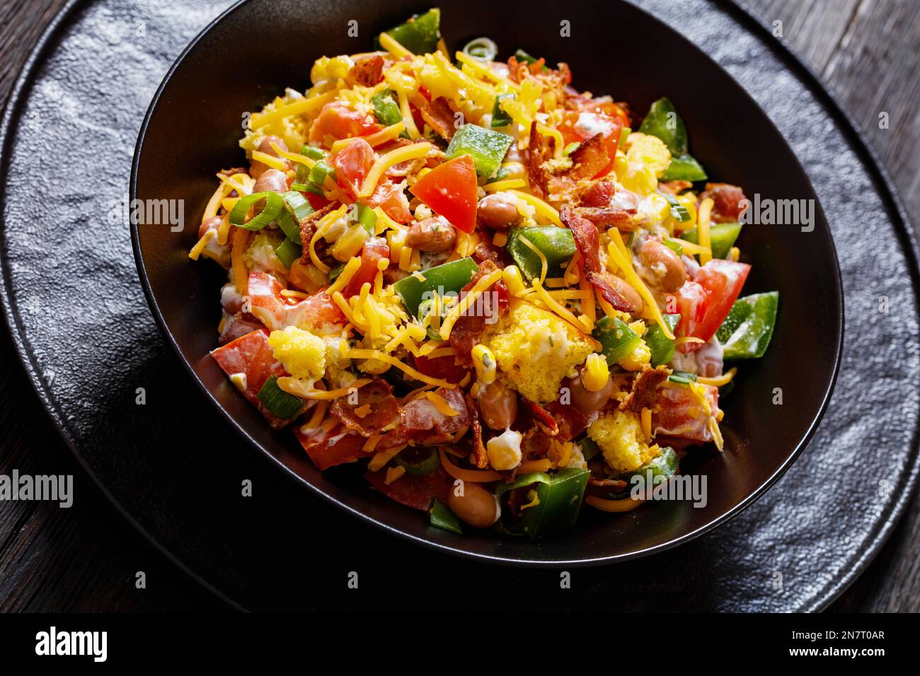 pinto Beans Speck Maisbrotsalat mit Tomaten, Paprika, Mais und Cheddar Käse in schwarzer Schüssel auf dunklem Holztisch, Nahaufnahme Stockfoto