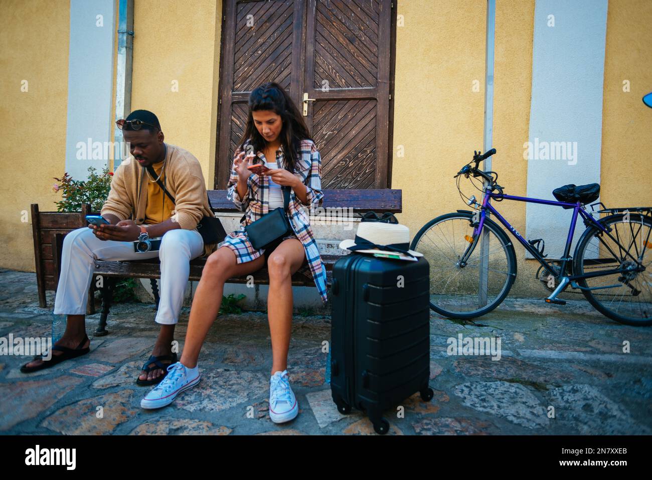 Wunderschöne multiethnische Freunde unterhalten sich auf ihren Telefonen, während sie eine Pause machen und auf einer Bank sitzen Stockfoto